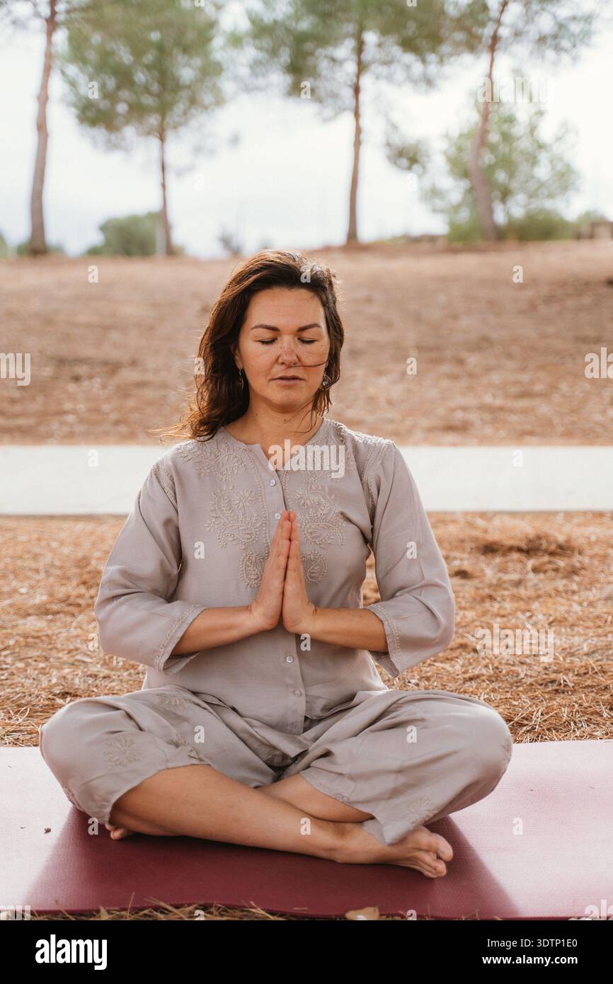 Donna seduta seduta a gambe incrociate all'aperto a meditare con gli occhi chiusi e le mani in posizione di preghiera. Circondato dalla natura, trasmette relax e mentalità Foto Stock