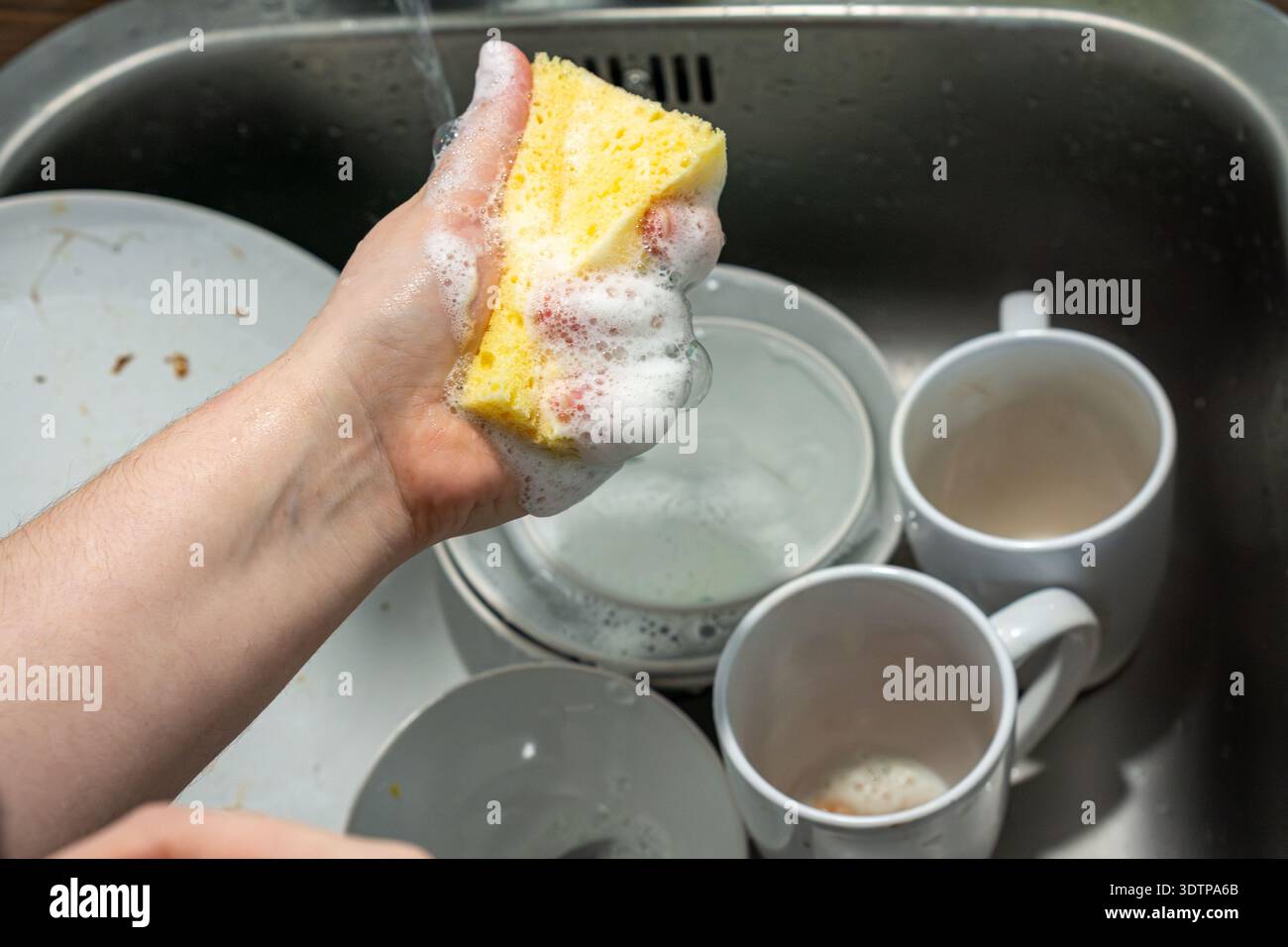 Primo piano di una mano che tiene una spugna gialla ricoperta di spessa schiuma di sapone durante il lavaggio dei piatti. Piastre e tazze sono visibili in un lavello in acciaio inox. Foto Stock