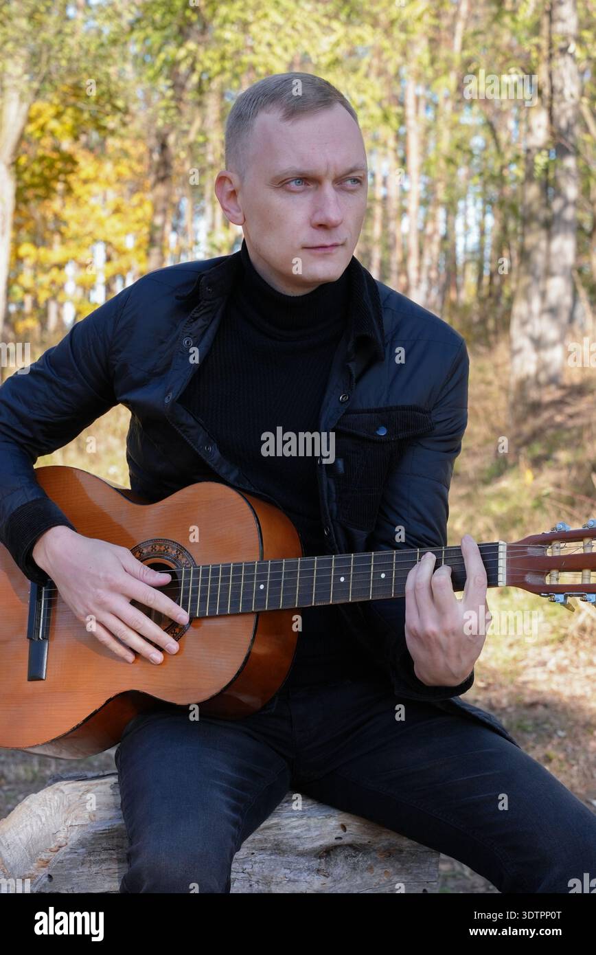 Bellissimo biondo che suona la chitarra acustica e canta rilassandosi all'aperto nella soleggiata giornata autunnale. Artista che impara e pratica a suonare strumenti musicali. L'autunno lascia il clima autunnale Foto Stock