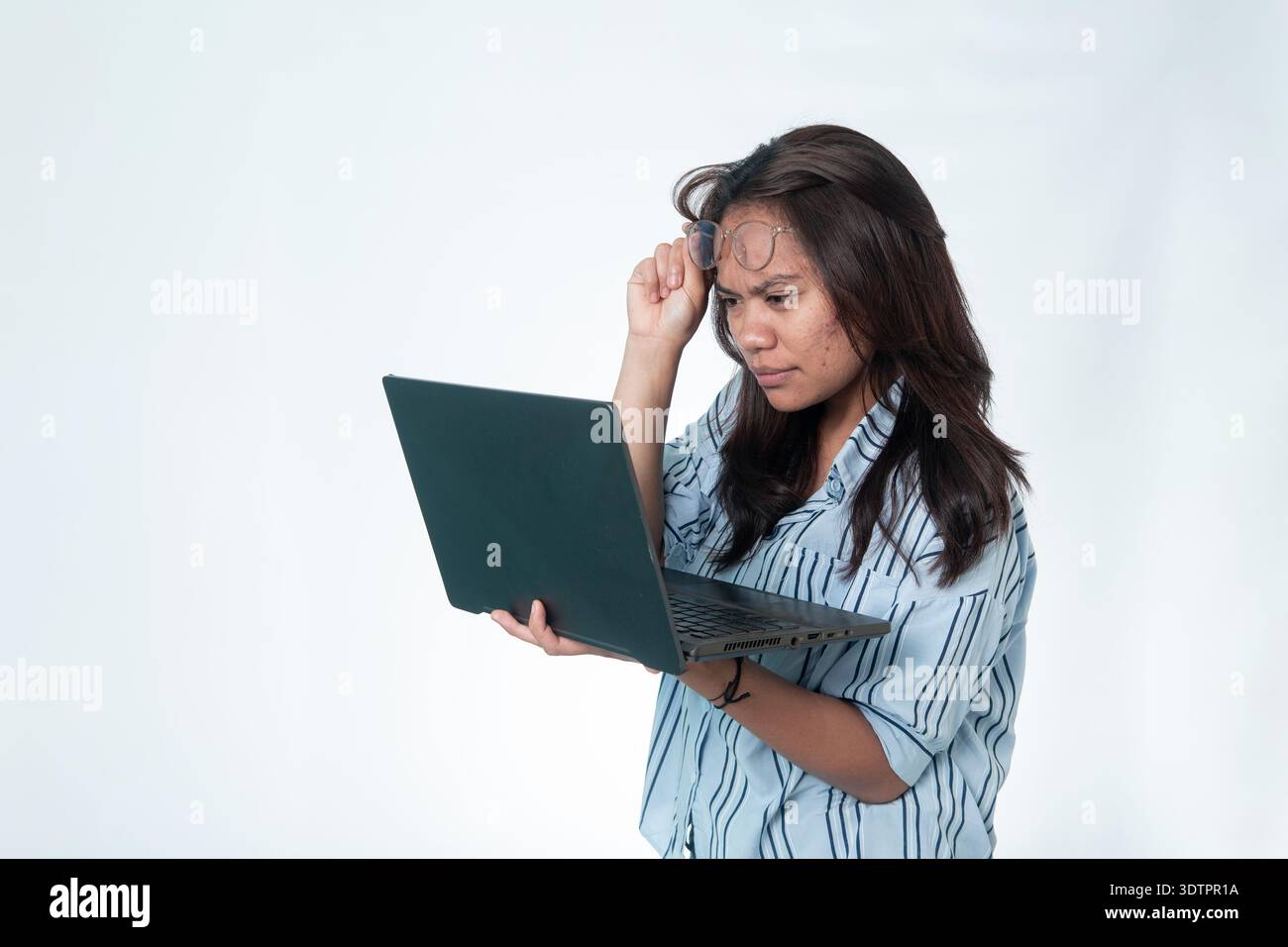 Donna indonesiana con una camicia a righe che solleva gli occhiali per guardare da vicino il suo laptop. Espressione analitica confusa o seria. Isolato su Who Foto Stock