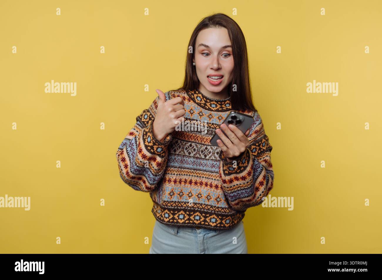 Una giovane donna interagisce con gioia con il suo smartphone, irrompendo di entusiasmo, mostrando il suo maglione colorato contro una parete gialla soleggiata, creando un Foto Stock
