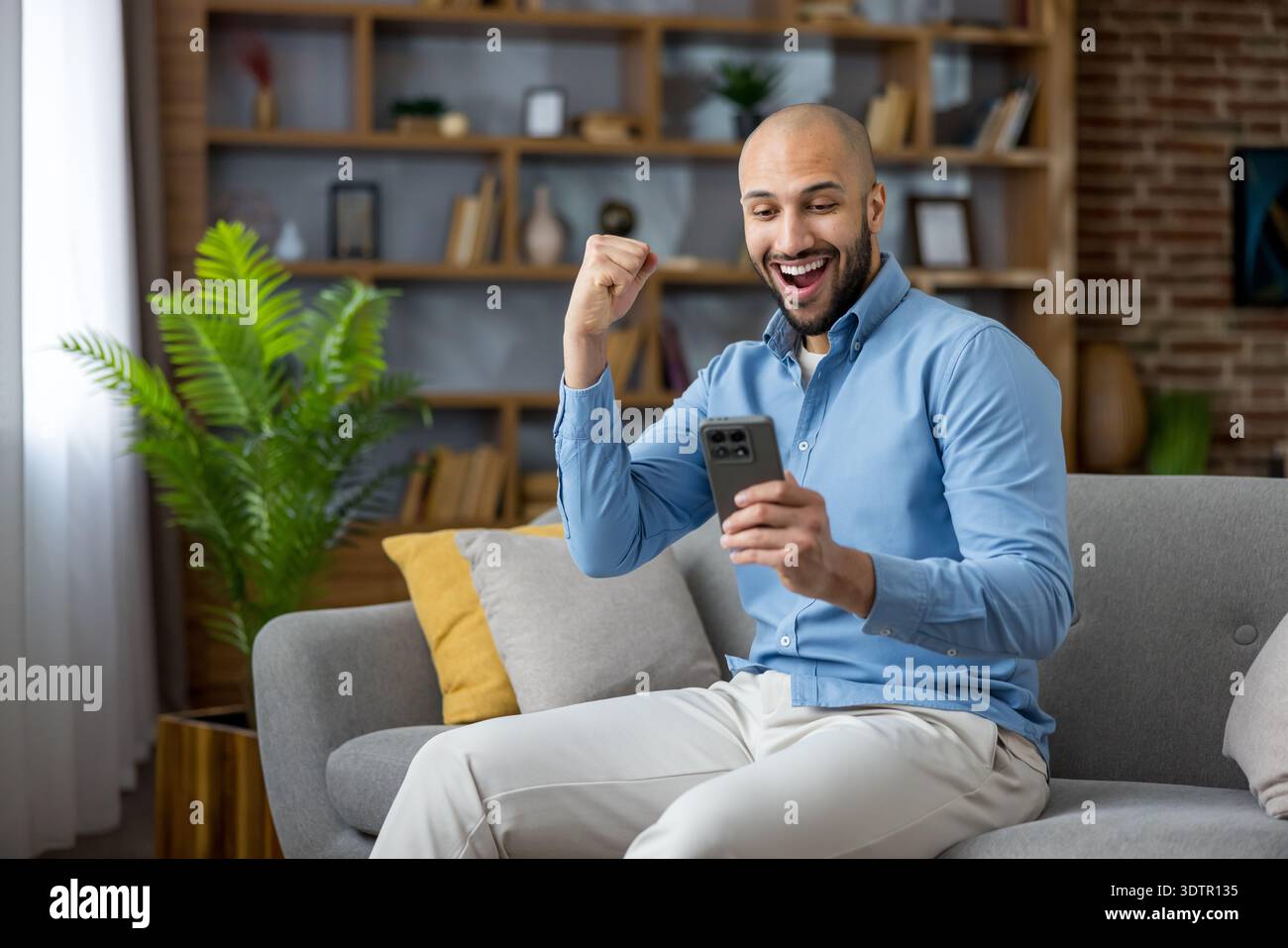 Un giovane uomo su un divano nel suo ufficio domestico celebra le buone notizie sul suo smartphone, sorridendo e tifa mentre controlla un messaggio, un'app o un aggiornamento sul mercato Foto Stock