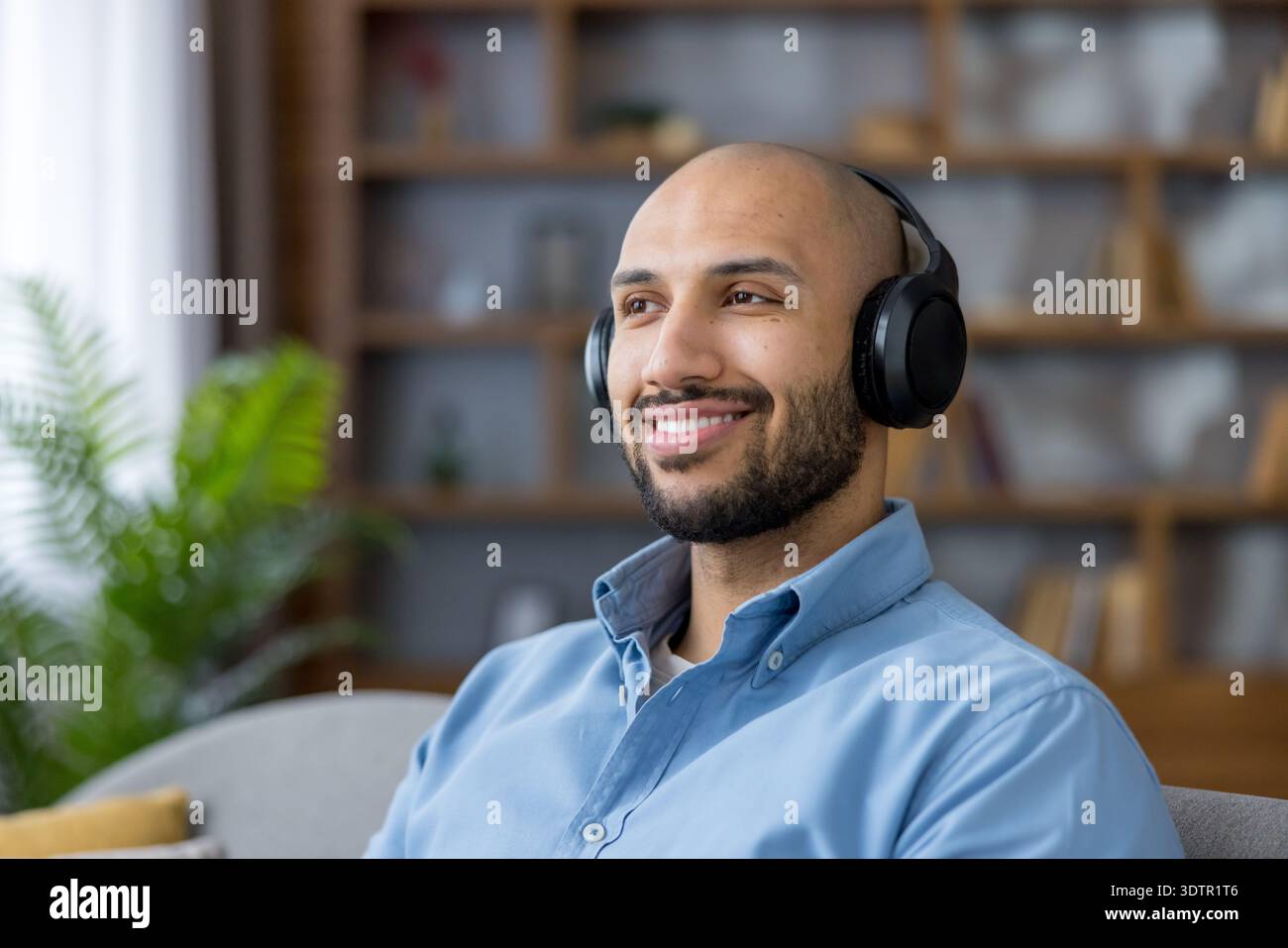 Giovane uomo che indossa una camicia blu e cuffie wireless nere, sorridendo felicemente mentre si rilassa e si gode l'audio digitale in un ambiente confortevole Foto Stock