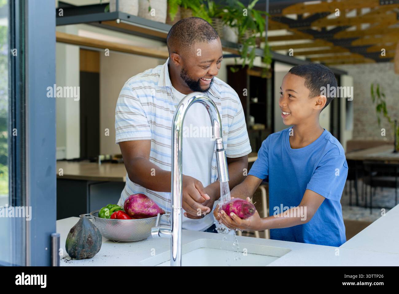 Padre e figlio africani che lavano il tubero viola sotto il rubinetto cromato al lavello della cucina, colino visibile Foto Stock