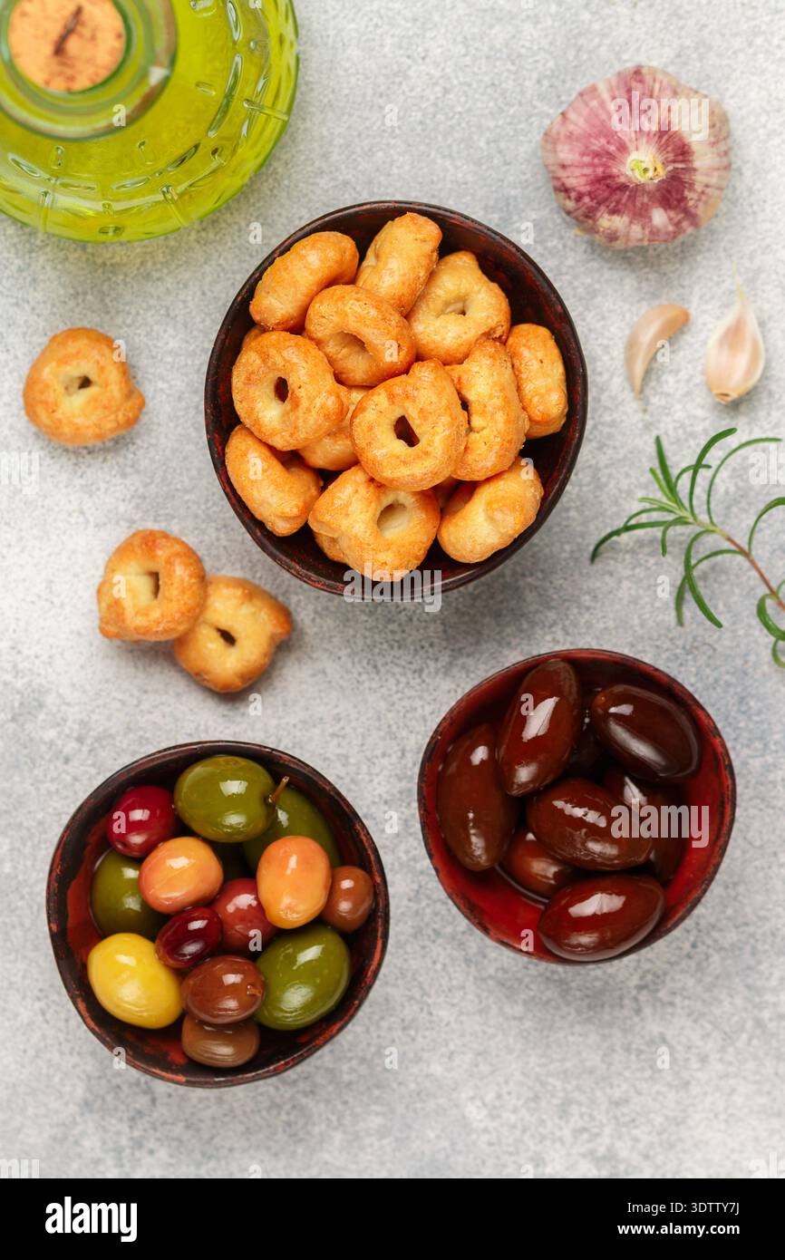 Tarallini: Piccoli bagel italiani tradizionali. Taralli. Snack gustoso. Olive nere e verdi, Kalamata, olio d'oliva, aglio e rosmarino su cemento grigio Foto Stock