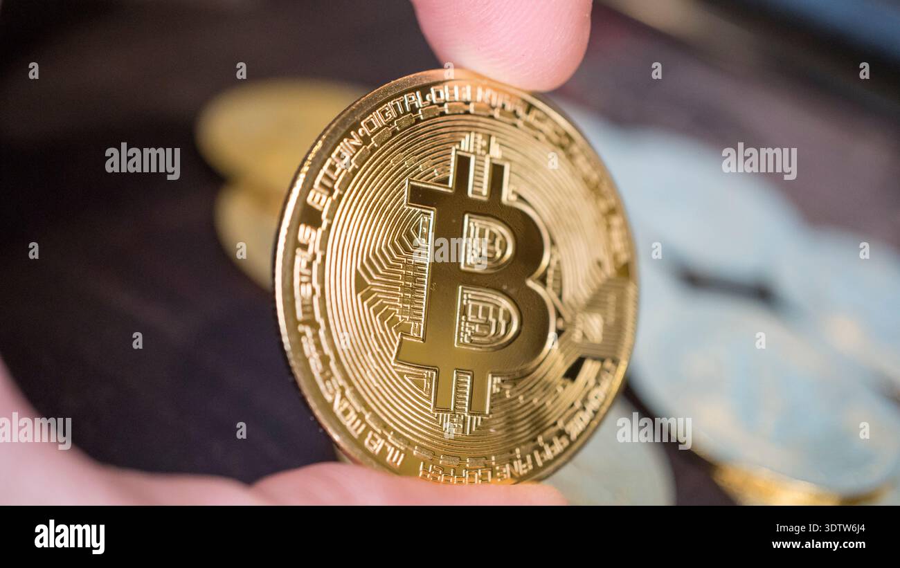 Primo piano di una mano che tiene in mano un bitcoin d'oro lucido con monete di criptovaluta sfocate sullo sfondo, simboleggiando la finanza digitale e gli investimenti. Foto Stock