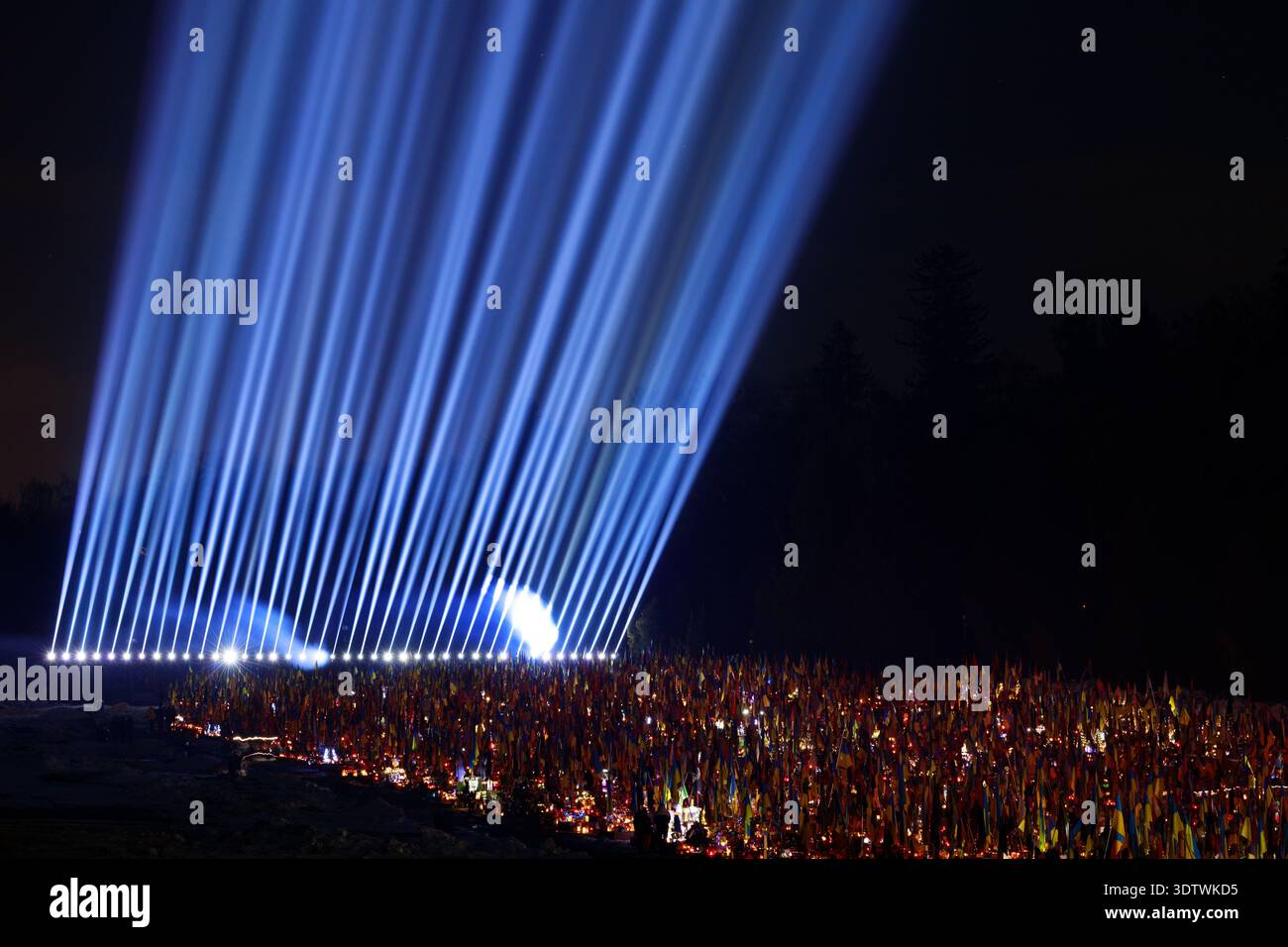 Un potente spettacolo di raggi di luce blu splende nel cielo notturno sopra un vasto campo pieno di bandiere ucraine, simboleggiando speranza e solidarietà Foto Stock