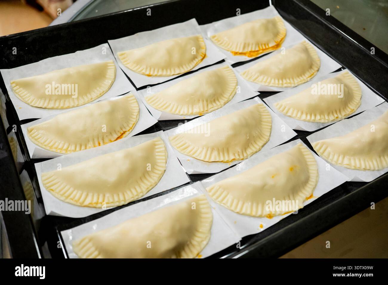 Una dozzina di empanadas crude preparate a casa sono disposte in modo ordinato su vassoi da forno rivestiti in pergamena, mostrando la loro perfetta forma a mezzaluna, pronte per la t Foto Stock
