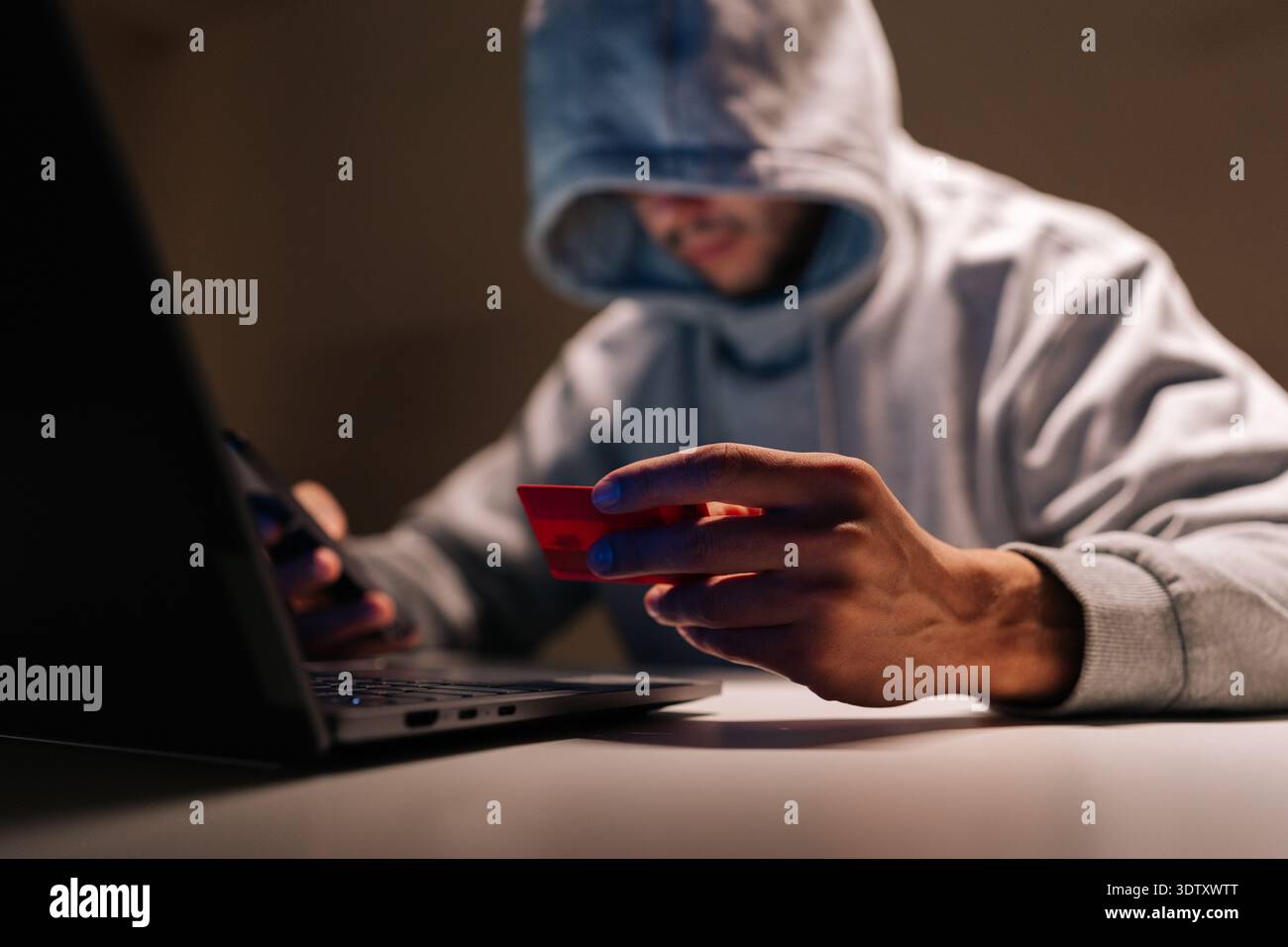Ritratto di una persona incappucciata e irriconoscibile in una stanza buia che tiene in mano il telefono e la carta di credito mentre utilizza il computer portatile, raffigurante frodi online, furti di identità e. Foto Stock