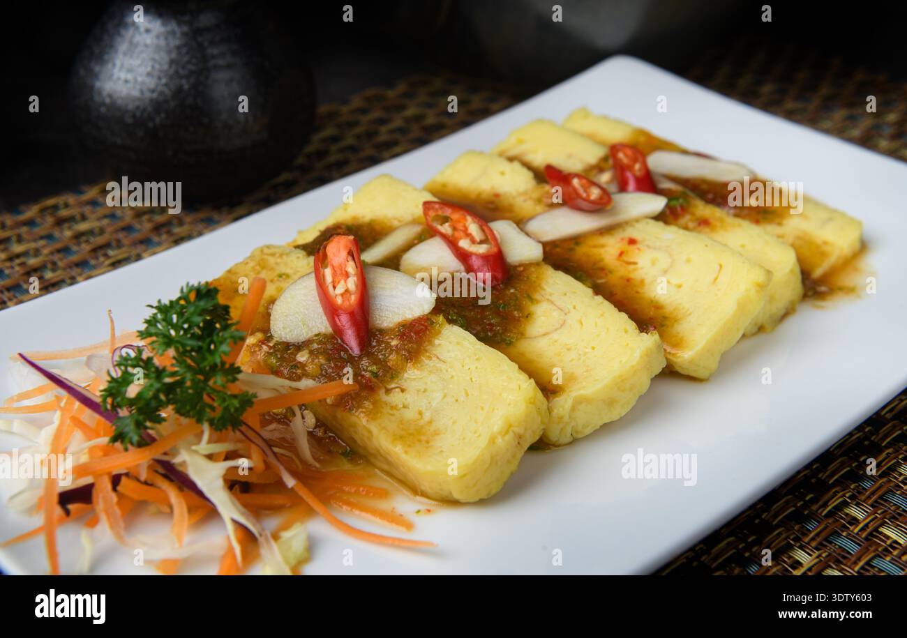 Fette di tamagoyaki giapponese (omelette dolci) condite con salsa piccante, disposte ordinatamente su un piatto bianco con verdure fresche. Foto Stock