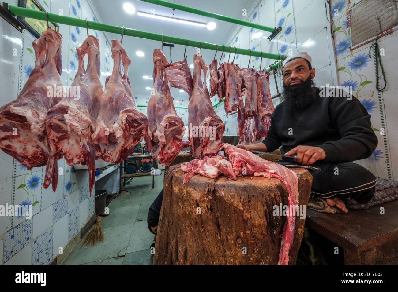 Un macellaio prepara tagli di carne su un grande blocco di legno in un negozio piastrellato con carcasse appese a un rotaia verde nella vecchia Delhi. Foto Stock