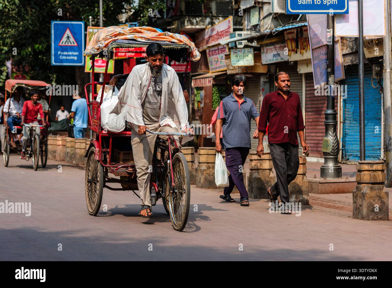 Un autista di risciò trasporta merci lungo Chandni Chowk, mentre i pedoni passano accanto a negozi e cartelli in lontananza nella vecchia Delhi. Foto Stock