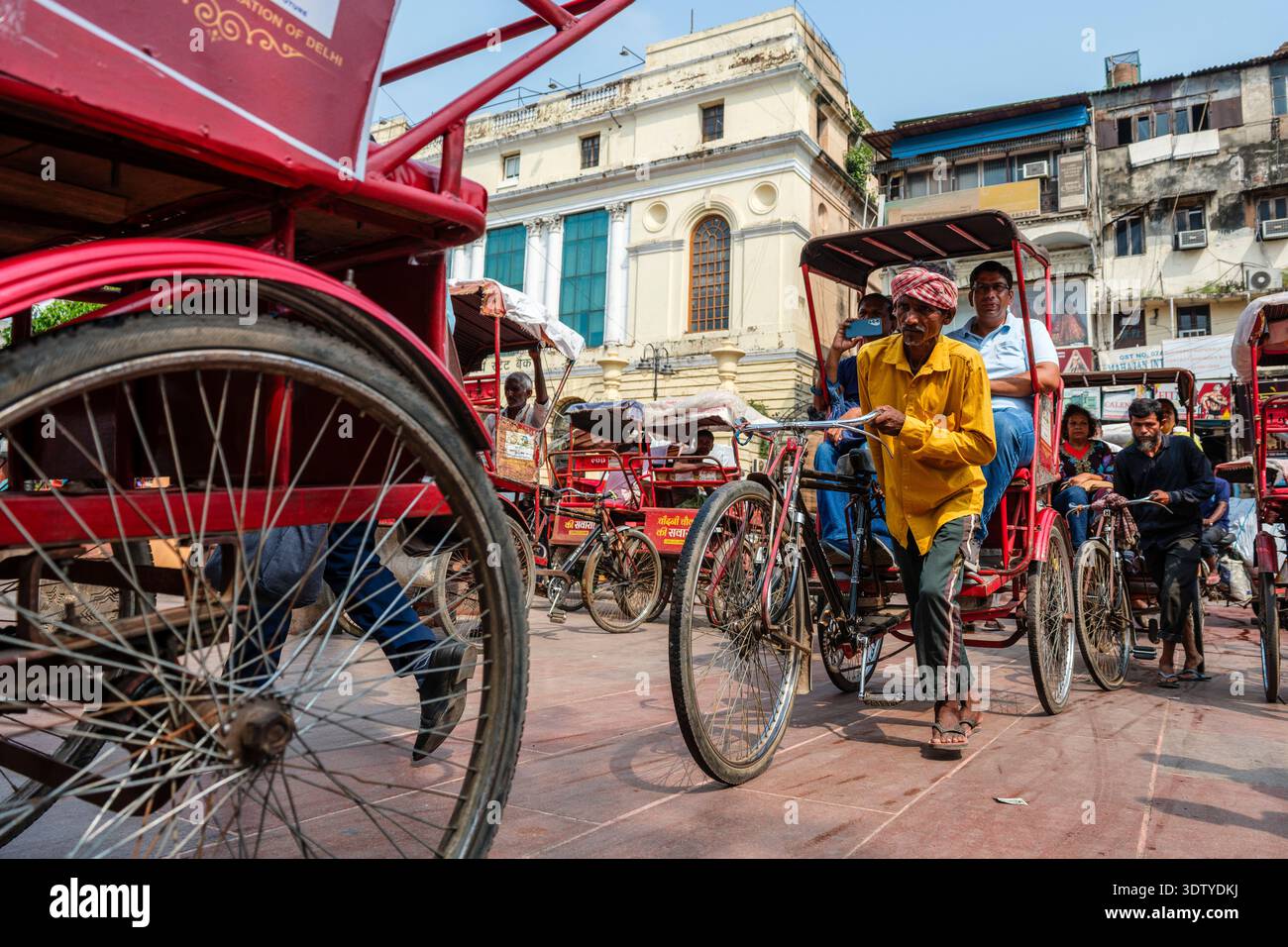 I risciò in bicicletta portano i passeggeri oltre lo storico edificio della State Bank of India lungo la trafficata via Chandni Chowk nella vecchia Delhi. Foto Stock