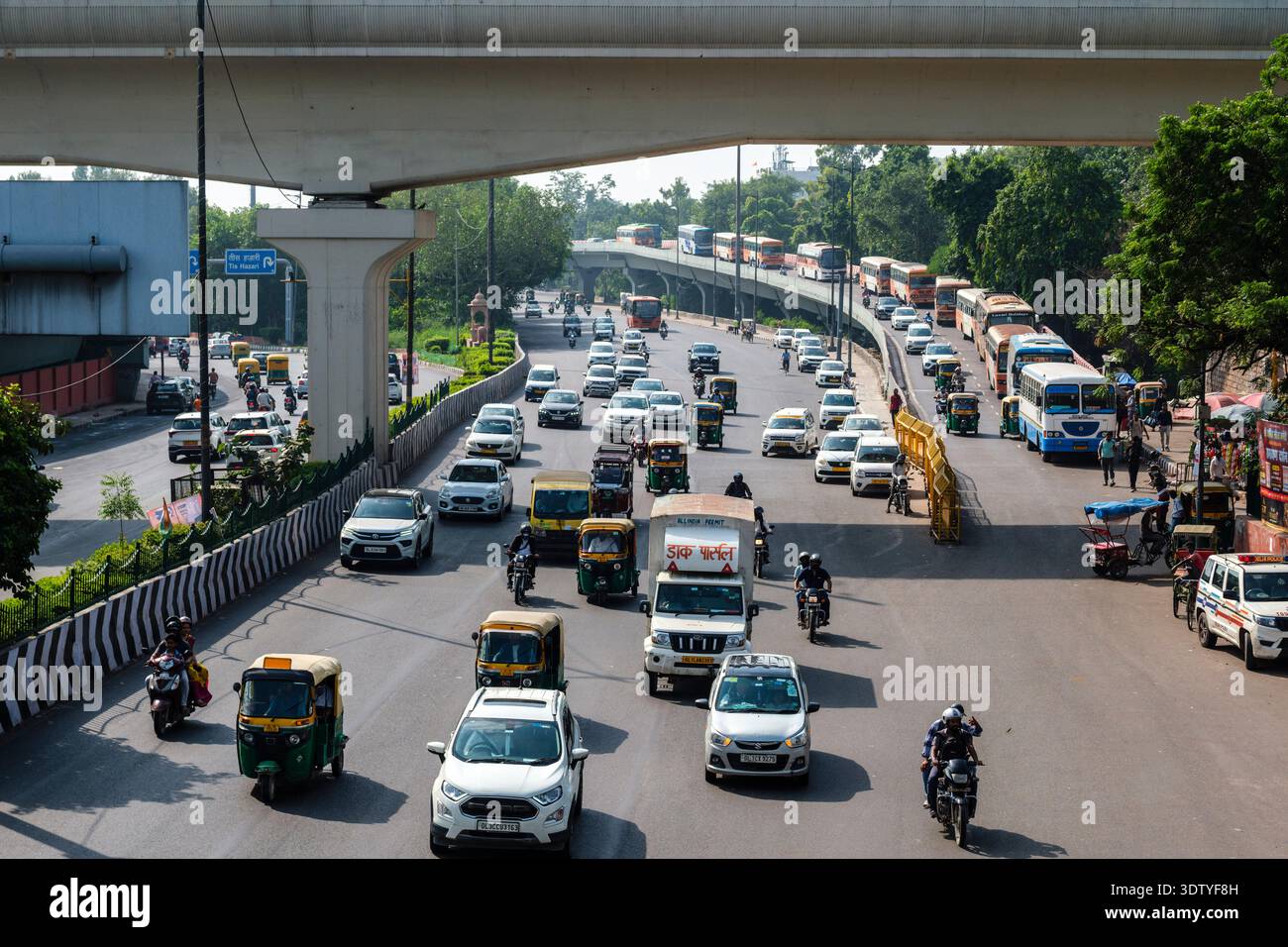 Il traffico intenso con auto, risciò e autobus scorre lungo una strada a più corsie sotto un viadotto della metropolitana in cemento nella vecchia Delhi. Foto Stock