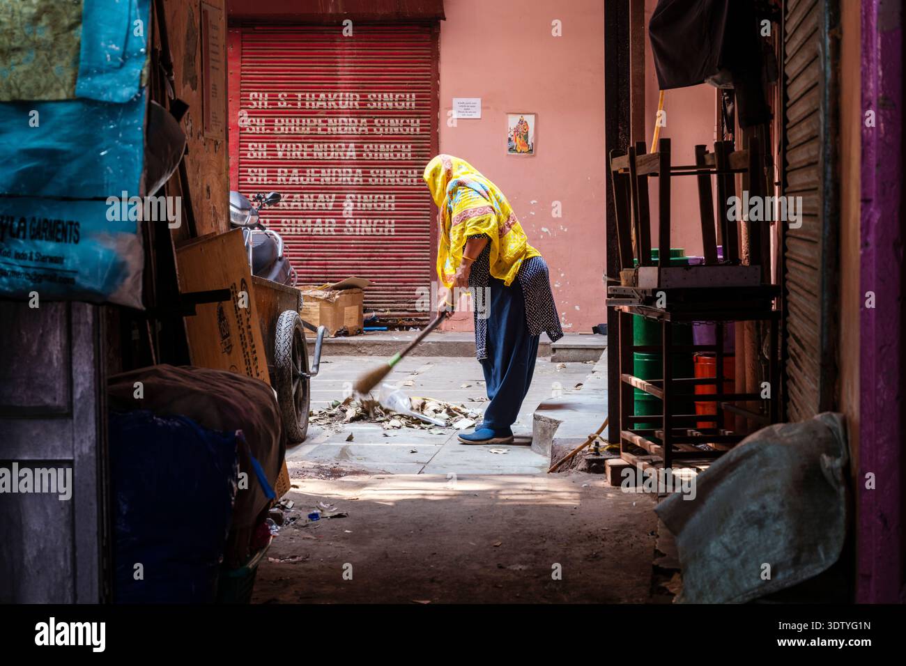 Una donna con un velo giallo spazia in uno stretto cortile con una scopa tradizionale vicino all'affollato Chandni Chowk nella Vecchia Delhi. Foto Stock