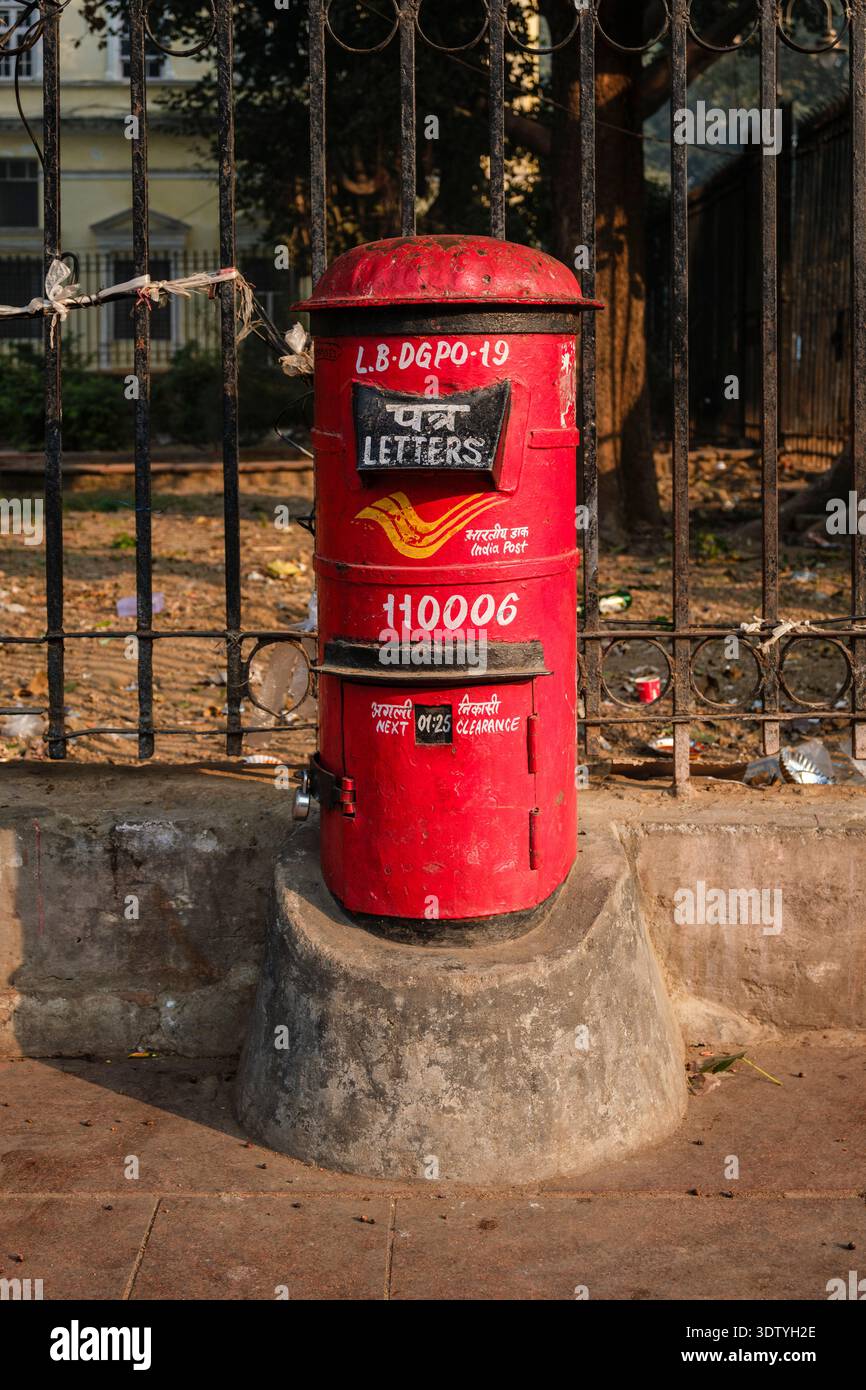 Casella di posta rossa dell'India su un marciapiede nella vecchia Delhi. Una recinzione di metallo nero e un edificio giallo pallido sono visibili in lontananza. Foto Stock