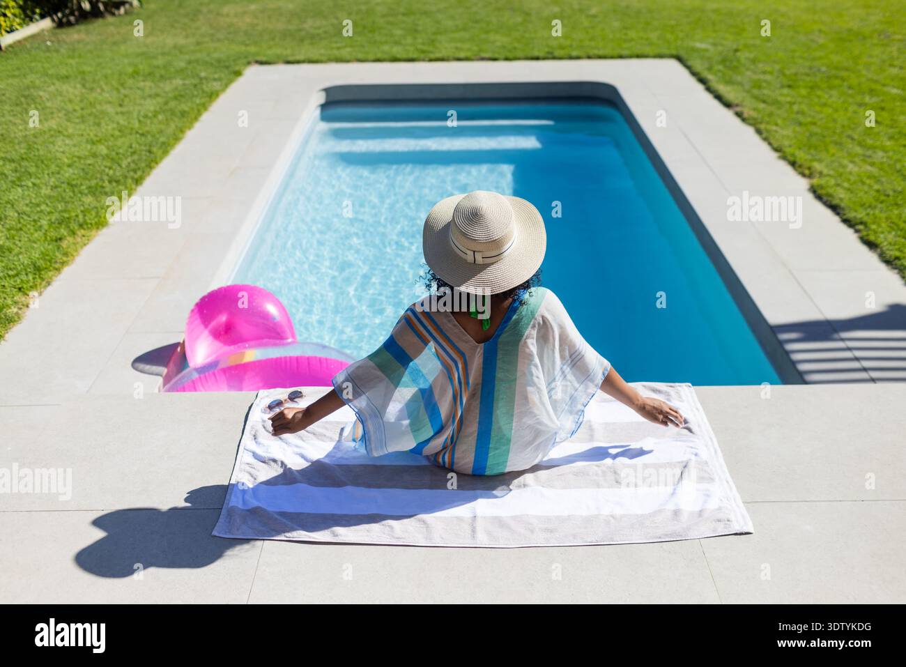 Donna seduta su un asciugamano bianco nella piscina del cortile con anello gonfiabile rosa che indossa un cappello di paglia Foto Stock
