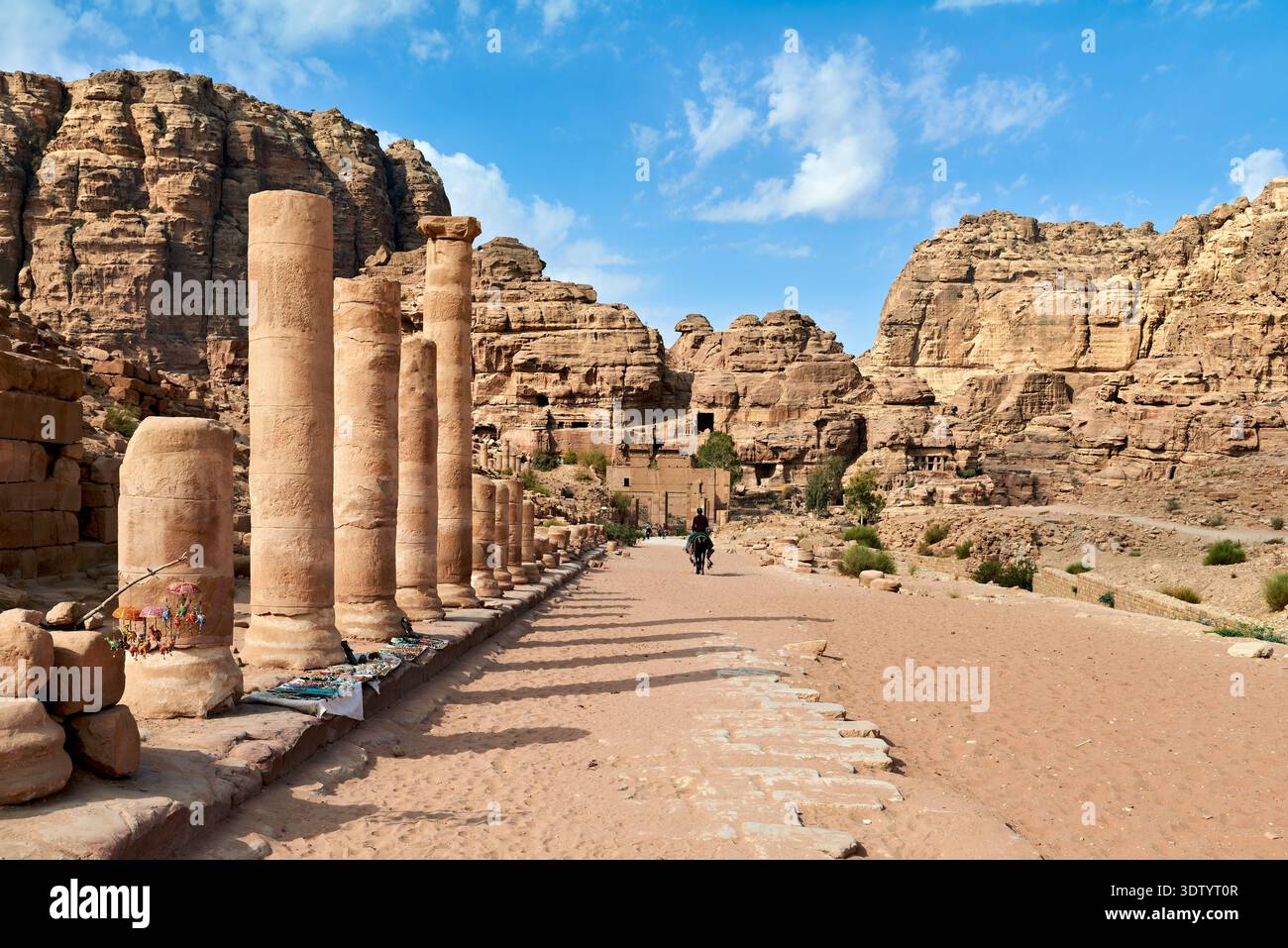 I turisti camminano attraverso la porta Adriana a Petra. L'area presenta colonne di pietra e scogliere rocciose sotto un cielo blu. Foto Stock