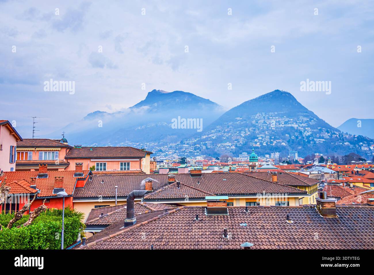 vista panoramica delle montagne dietro i tetti storici urbani di Lugano, Svizzera Foto Stock