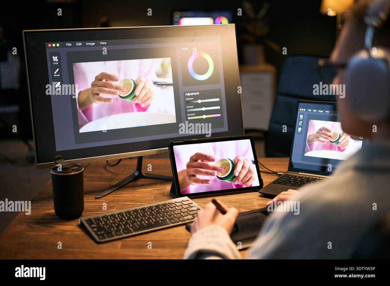 Giovane uomo caucasico che utilizza un tablet grafico e uno stilo per l'editing di foto di prodotto su computer e tablet, indossa le cuffie, concentrandosi sul processo di ritocco digitale alla scrivania Foto Stock