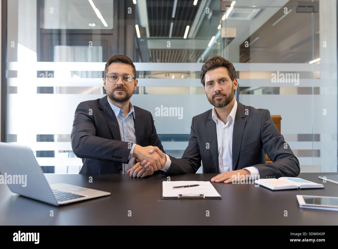 Due uomini d'affari fiduciosi che stringono la mano a una scrivania con un laptop e documenti, simboleggiando un accordo di partnership di successo o il completamento di un accordo professionale in un ufficio moderno Foto Stock