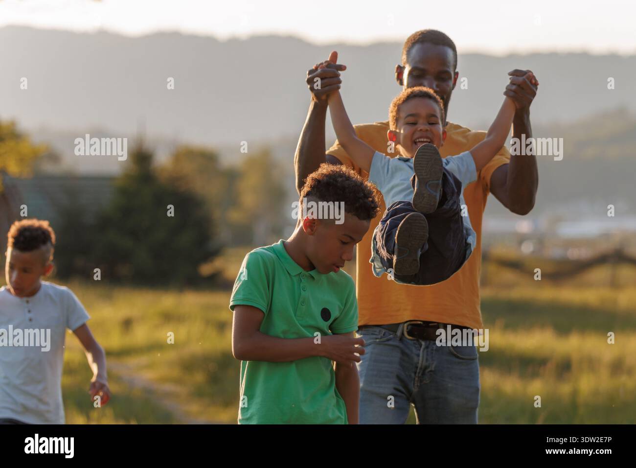 Padre e due bambini condividono risate e giocano all'aperto mentre il sole tramonta sul paesaggio creando momenti felici insieme. Foto Stock