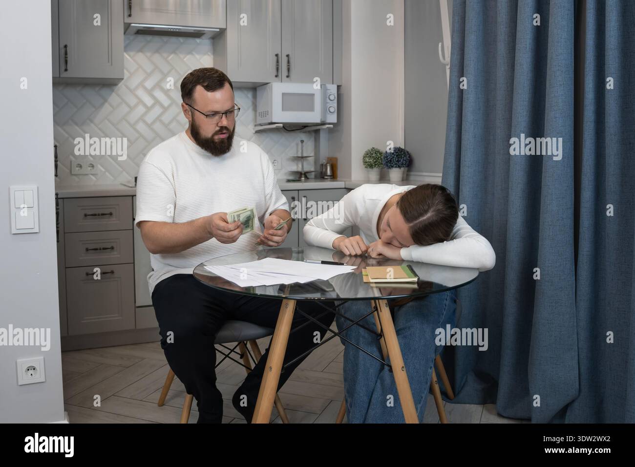 Coppia che affronta difficoltà finanziarie in cucina, con l'uomo che conta i soldi e la donna che sembra esausta, simboleggiando stress, debito e budget Foto Stock