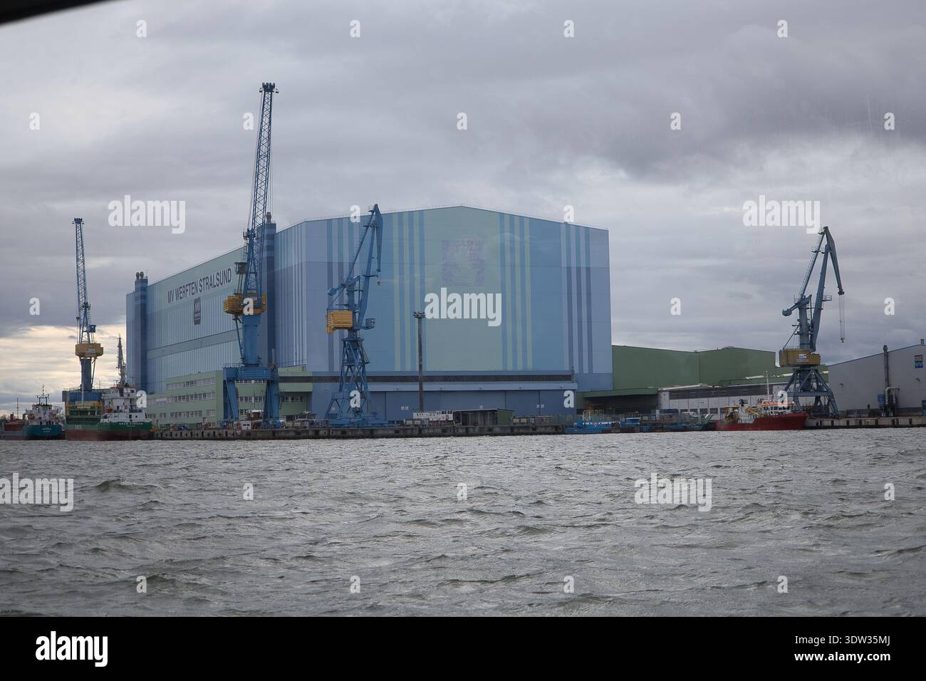 Vista dettagliata del cantiere navale MV Werften Stralsund con gru a cavalletto blu e una nave da carico presso il molo di Strelasund Foto Stock