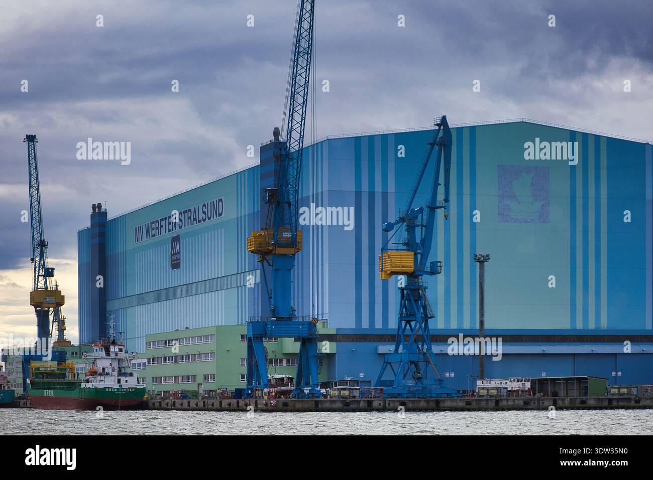Vista dettagliata del cantiere navale MV Werften Stralsund con gru a cavalletto blu e una nave da carico presso il molo di Strelasund Foto Stock