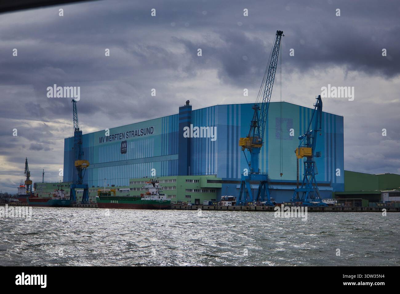 Grande sala di costruzione navale di MV Werften Stralsund con gru a cavalletto sulle rive dello Strelasund sotto un cielo nuvoloso Foto Stock