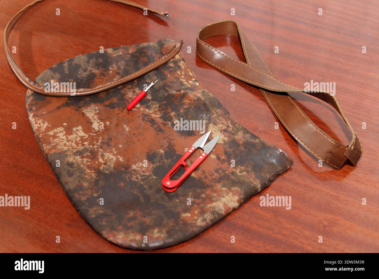 Un sacchetto usurato con motivi marroni viene smontato per la riparazione su un tavolo di legno. Cesoie per filetti con manico rosso e ripper per cuciture appoggiati sulla parte superiore, pronti per essere cuciti o upc Foto Stock
