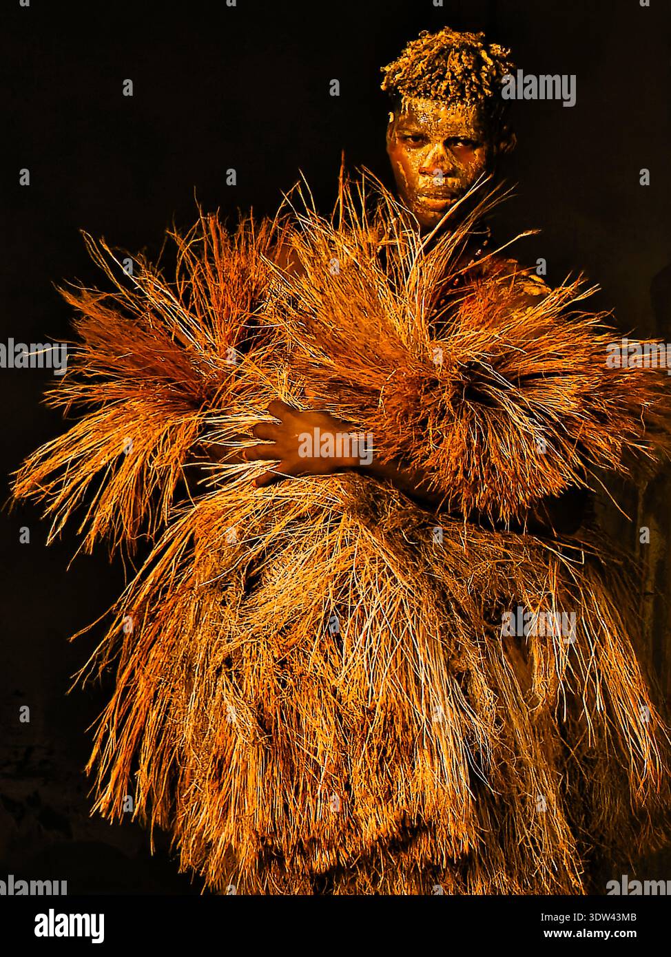 Una persona con un tradizionale costume di paglia si erge su uno sfondo scuro. Foto Stock