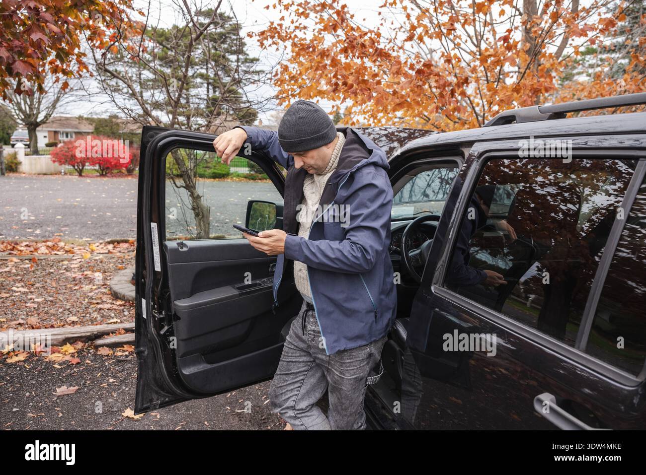Uomo in piedi davanti alla porta dell'auto aperta e che controlla lo smartphone all'aperto in autunno Foto Stock