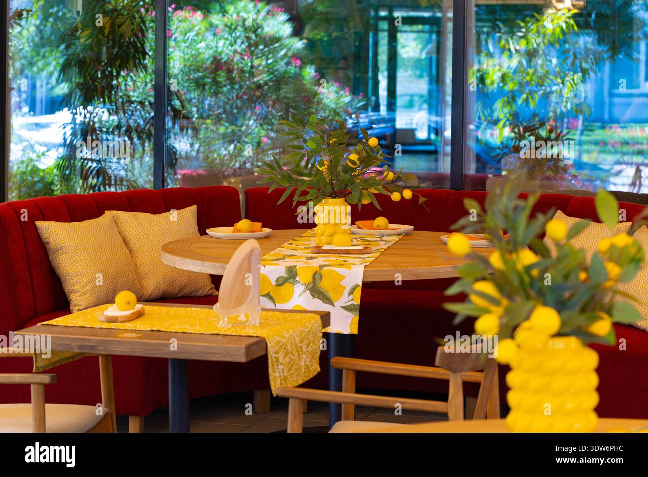 Un vivace ristorante interno presenta un tavolo rotondo in legno adornato con motivi al limone e vasi gialli, completato da lussuosi tavoli rossi a banchetto Foto Stock