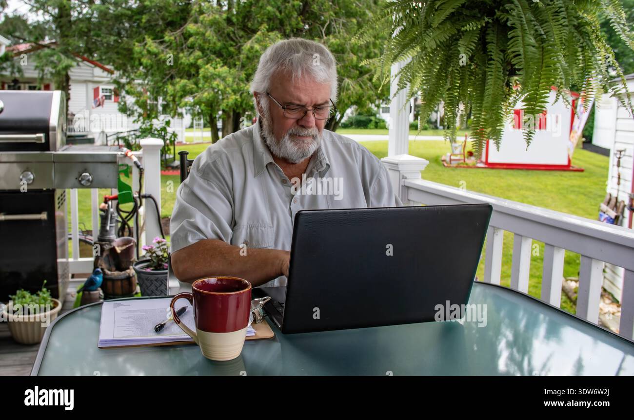 Un uomo siede a un tavolo su un portico, lavora su un computer portatile mentre sorseggia un caffè. Ha i capelli bianchi del viso e indossa una camicia leggera. Il cortile sul retro è verde con alberi e un grill nelle vicinanze. Foto Stock