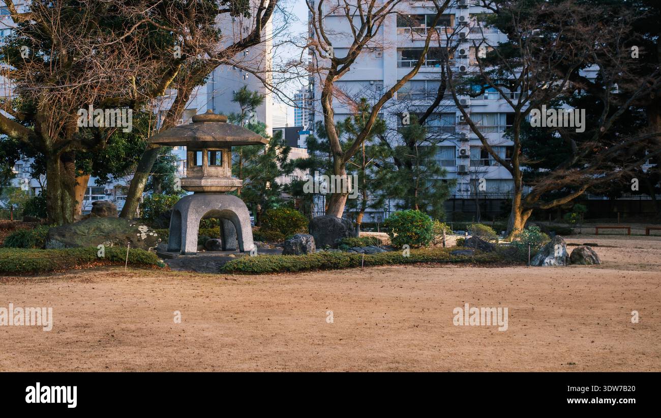 Una tradizionale lanterna in pietra sorge nel giardino paesaggistico di Kyū-Iwasaki-tei, incorniciata da alberi invernali e da edifici moderni che si riflettono Foto Stock