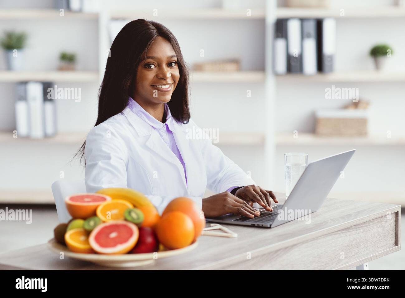 Nutrizionista che lavora su un laptop con frutta davanti presso Bright Office Space Foto Stock