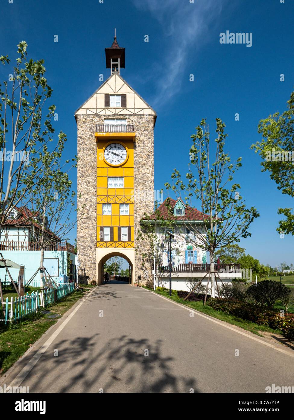 Immagine verticale di un'alta torre dell'orologio gialla con un cancello ad arco, che funge da ingresso simbolo in un villaggio in stile europeo o parco a tema. Foto Stock