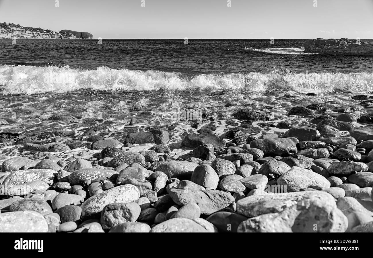 Paesaggio monocromatico di una spiaggia di ciottoli con onde infranti e schiuma marina a Cala Llobella, Benissa, Alicante, Spagna. Bianco e nero ad alto contrasto Foto Stock