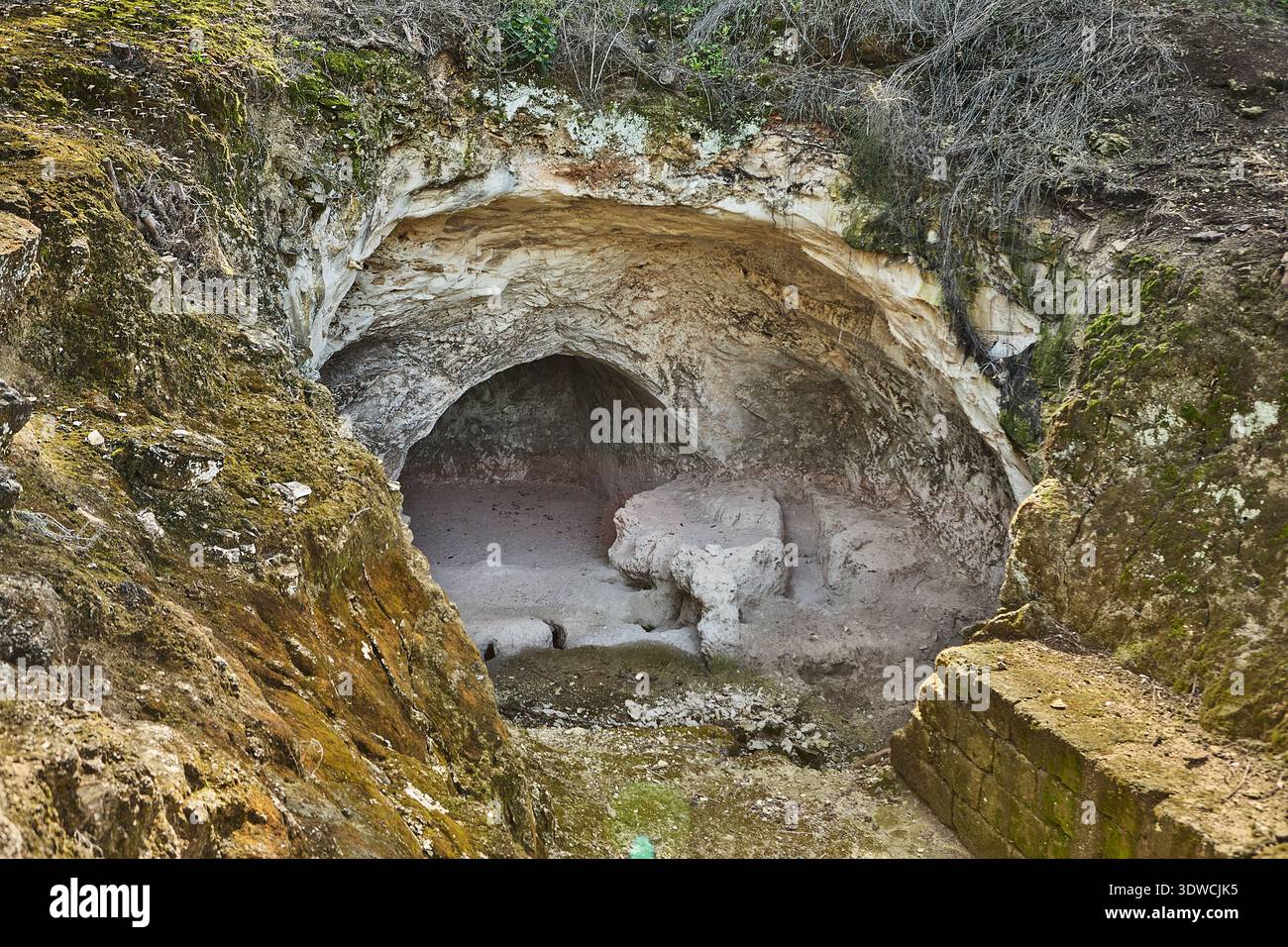 Ingresso naturale nella grotta con pareti rocciose e vegetazione ricoperta, che conduce ad un interno buio, circondato da terreno terroso e scalini in pietra in una zona rurale Foto Stock