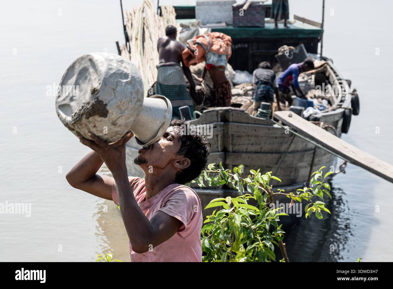 Satkhira, Bangladesh - 21 ottobre 2025: Vista di un uomo che beve da un grande container vicino a una barca, che riflette la vita quotidiana lungo le rive del fiume. Foto Stock
