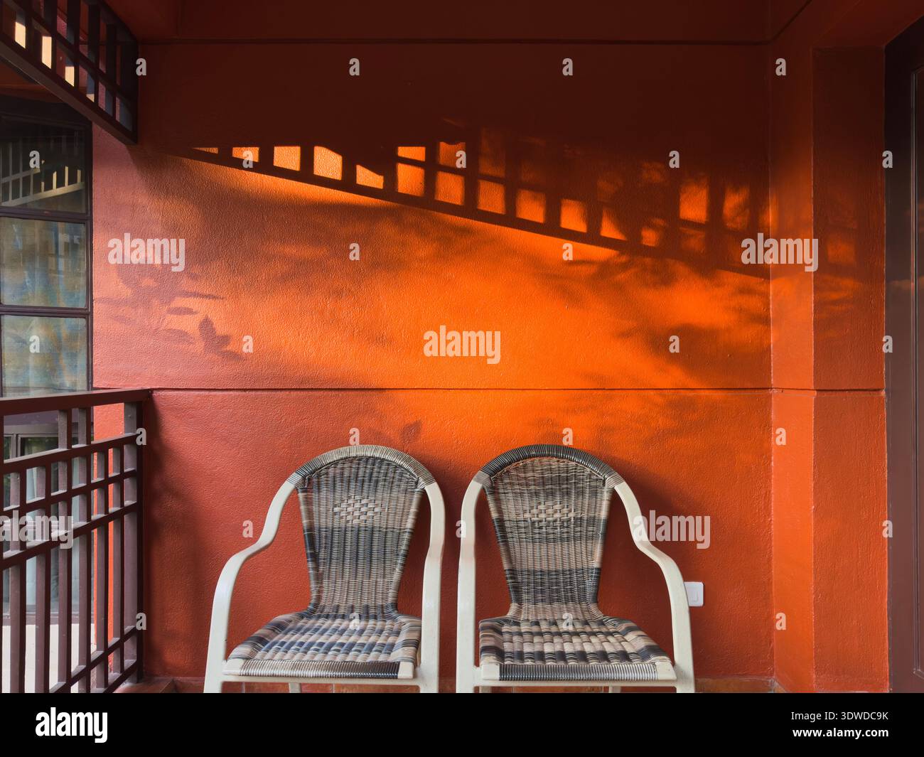 Due sedie in rattan con cuscini bianchi su un angolo murale rosso-arancio testurizzato, spettacolari ombre allungate dalle spranghe della finestra Foto Stock