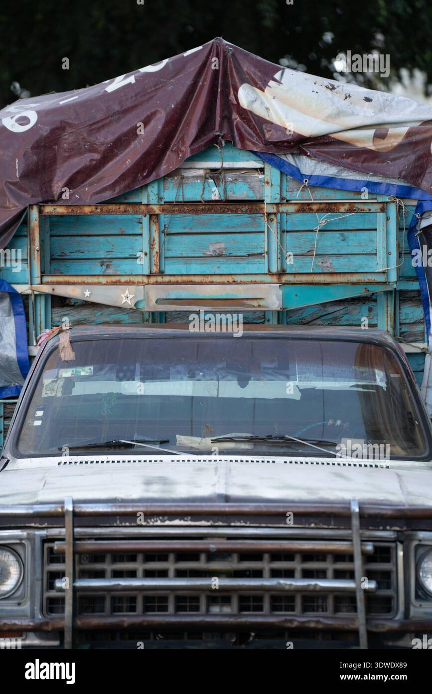 Un veicolo in condizioni atmosferiche con una griglia anteriore arrugginita è parcheggiato, parzialmente coperto da un telone marrone. Lo sfondo di legno blu aggiunge un rustico e vintage f Foto Stock