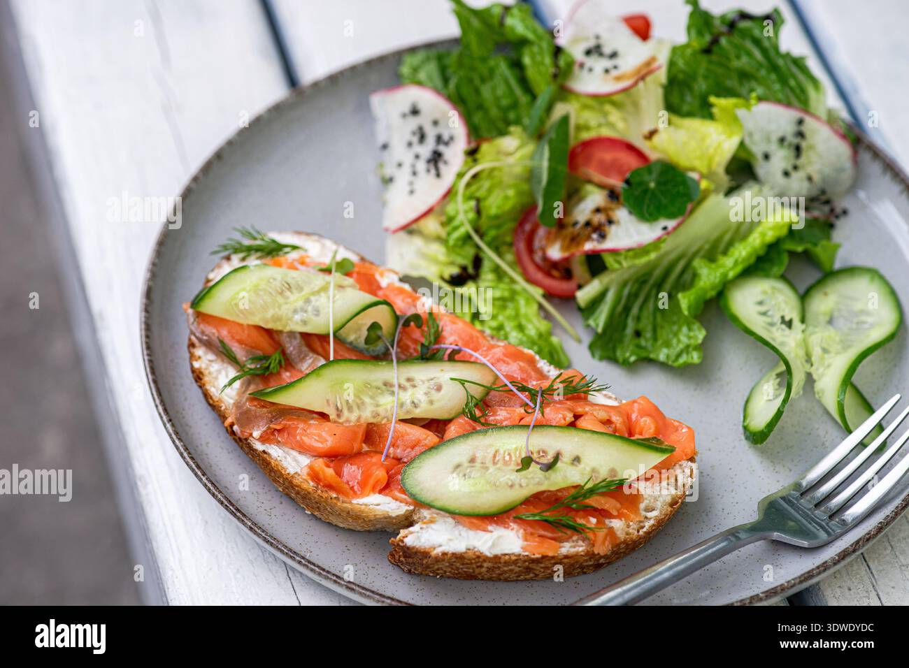Delizioso toast al salmone con fette di cetriolo e micro erbe aromatiche, accompagnato da un'insalata fresca con lattuga, pomodori, ravanelli e semi di sesamo Foto Stock