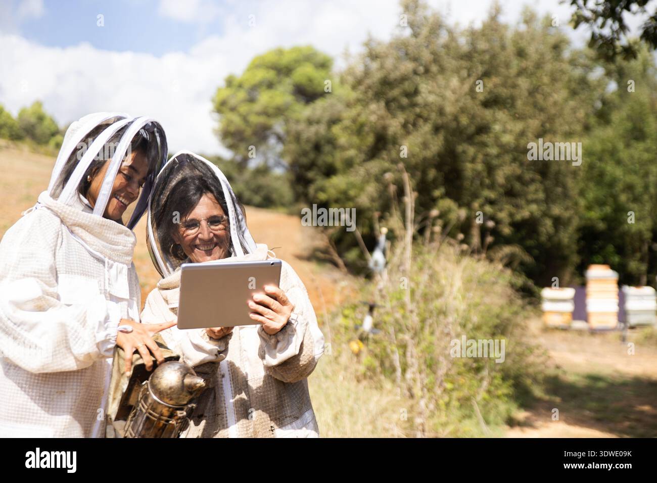 Due donne in tute protettive studiano le informazioni sull'alveare su una tavoletta, evidenziando le pratiche di apicoltura e il ruolo delle api mellifere nella natura e nel benessere. Foto Stock