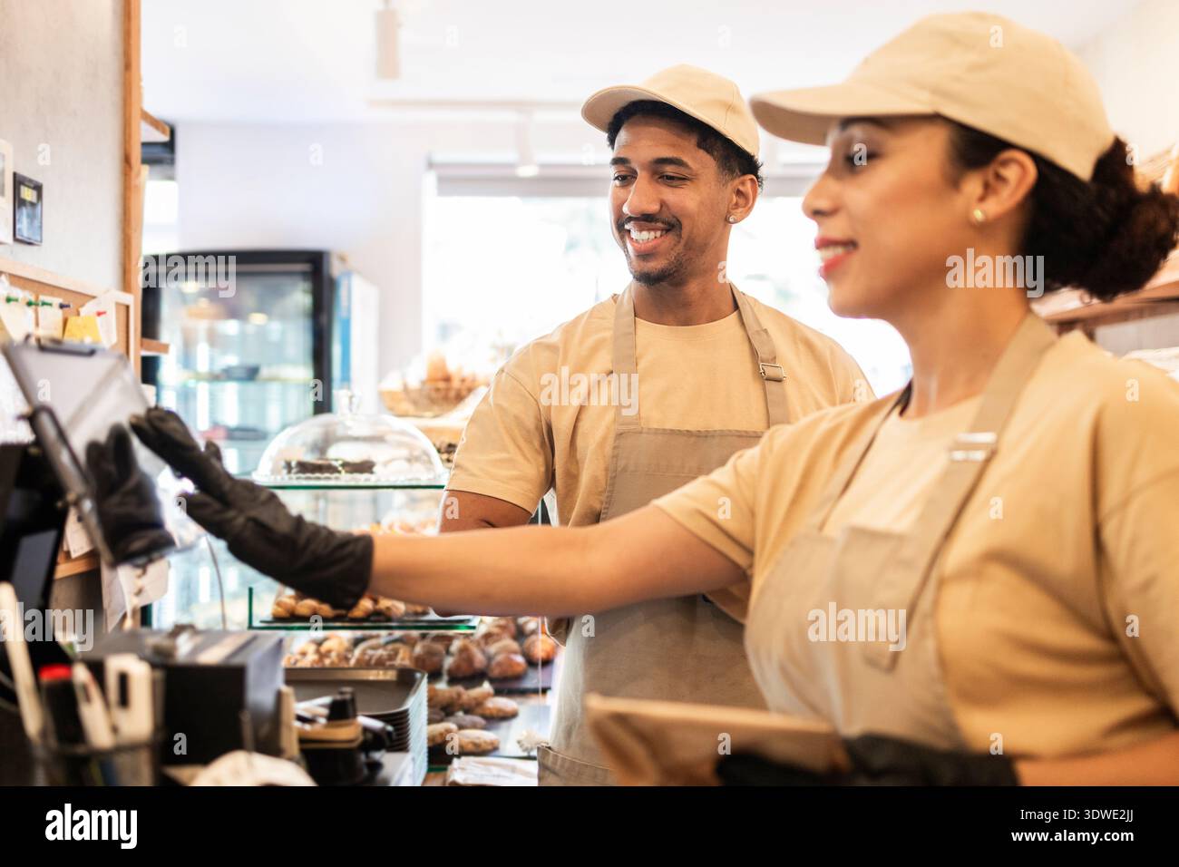 Due lavoratori del caffè, un uomo e una donna, collaborano allegramente alla loro panetteria. Sorridono mentre gestiscono le attività in un ambiente accogliente pieno di g al forno Foto Stock
