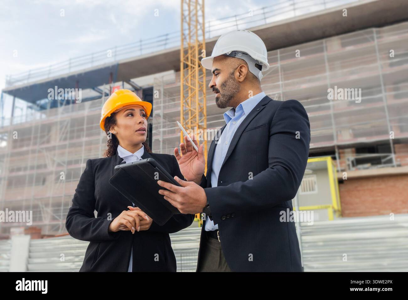 Due ingegneri con elmetti discutono di un progetto di costruzione in loco. Hanno un tablet e documenti, collaborando allo sviluppo di edifici in un ambiente urbano Foto Stock