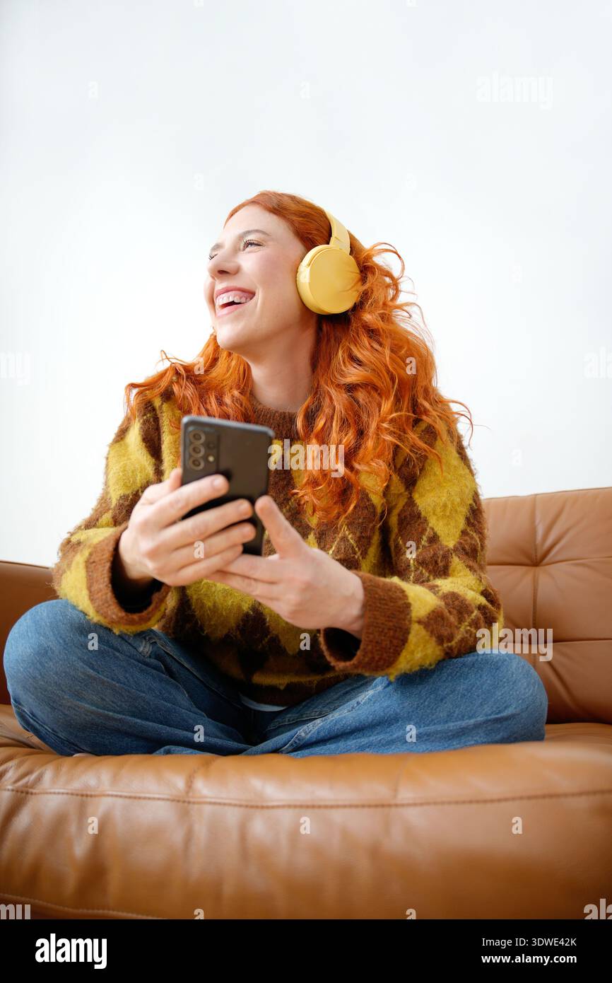 Donna con i capelli rossi che indossa le cuffie gialle, si siede a gambe incrociate su un divano in pelle marrone, sorridendo gioiosamente tenendo lo smartphone in entrambe le mani, e. Foto Stock