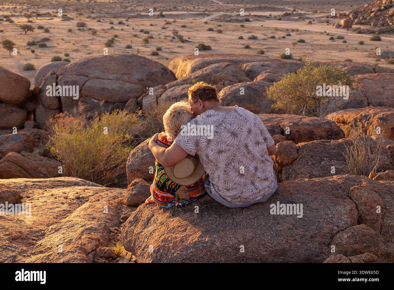 Una coppia si abbraccia su un terreno roccioso, ammirando uno splendido tramonto sull'ampio deserto della Namibia. Le calde sfumature del sole creano una serena, r Foto Stock