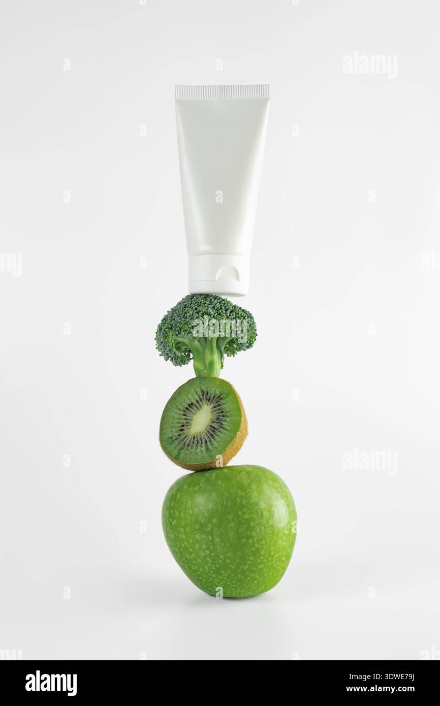 Immagine di un tubo per la crema cosmetica bilanciato con mela verde, kiwi e broccoli, che simboleggia l'uso di ingredienti alimentari biologici nei prodotti di bellezza per h Foto Stock